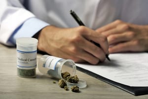 image of the hands of a person in a lab coat writing on papers that are sitting next to an open bottle of medical marijuana