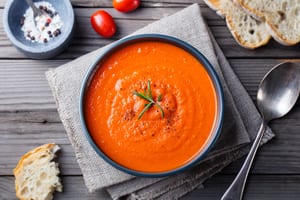 image of bowl of tomato soup with small garnish on top and a napkin underneath the bowl, and a spoon and bread next to the bowl