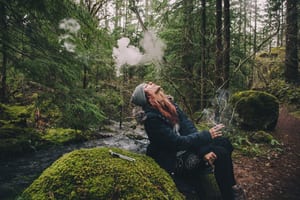 image of a woman sitting on a rock in the woods, smoking weed and blowing the smoke up above her head, showing how you can avoid marijuana red eyes by staying away from smoke