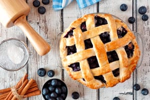 image of blueberry pie with a lattice top and a rolling pin, blueberries, and cinnamon partially out of frame
