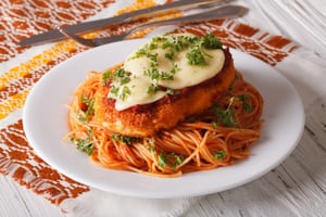 photo of chicken parmesan on a white plate, the pasta has red sauce and the breaded chicken has mozzarella on it with garnish on top