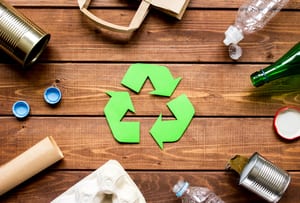a recycling symbol in the center of a wood table with recyclable items around it like water bottles, glass bottle, a paper towel roll