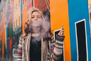 image of a blonde woman standing in front of a mural holding a vape and blowing smoke out of her mouth