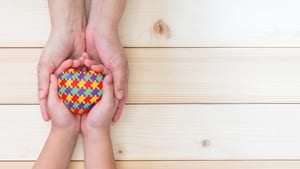 image of two hands reaching out over a wood table to hold a heart with the autism design on puzzle pieces